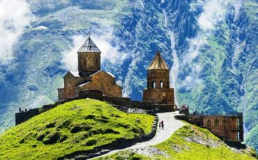 slika cerkve in dveh zvonikov na vrhu zelene planine, v ozadju strmo pobočje hribov v Gruziji