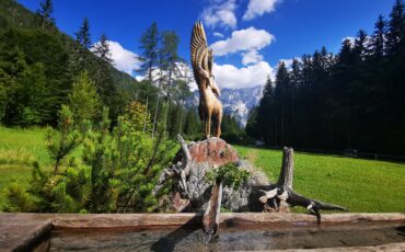 lesena skulptura kozice in price ujede, na skali, v ozadju zeleni travnik, gozd in gore