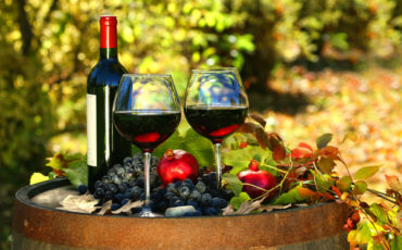 dva vinska kozarca z rdečim vinom, steklenica vina, temno grozdje in dekoracija, postavljena na vinski sod, v ozadju vinska trta