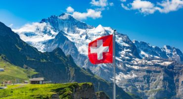 Swiss-flag-waving-and-tourists-admire-the-peaks-of-Monch-and-Jungfrau-mountains-on-a-Mannlichen-viewpoint-Bernese-Oberland-Switzerlandshutterstock_1467524654